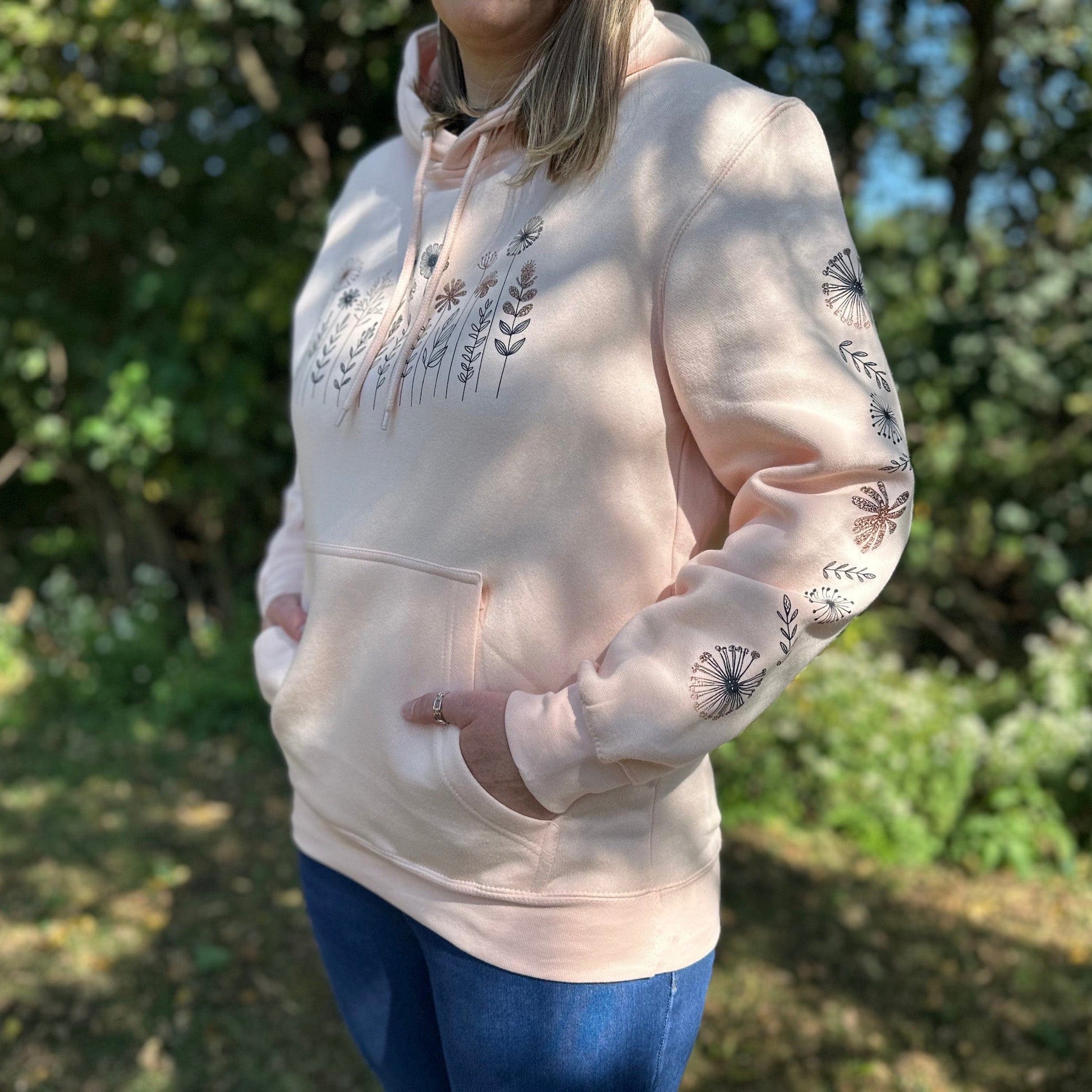 Woman standing in the woods wearing a pale pink hoodie that has a flower design on the chest and down the sleeve.