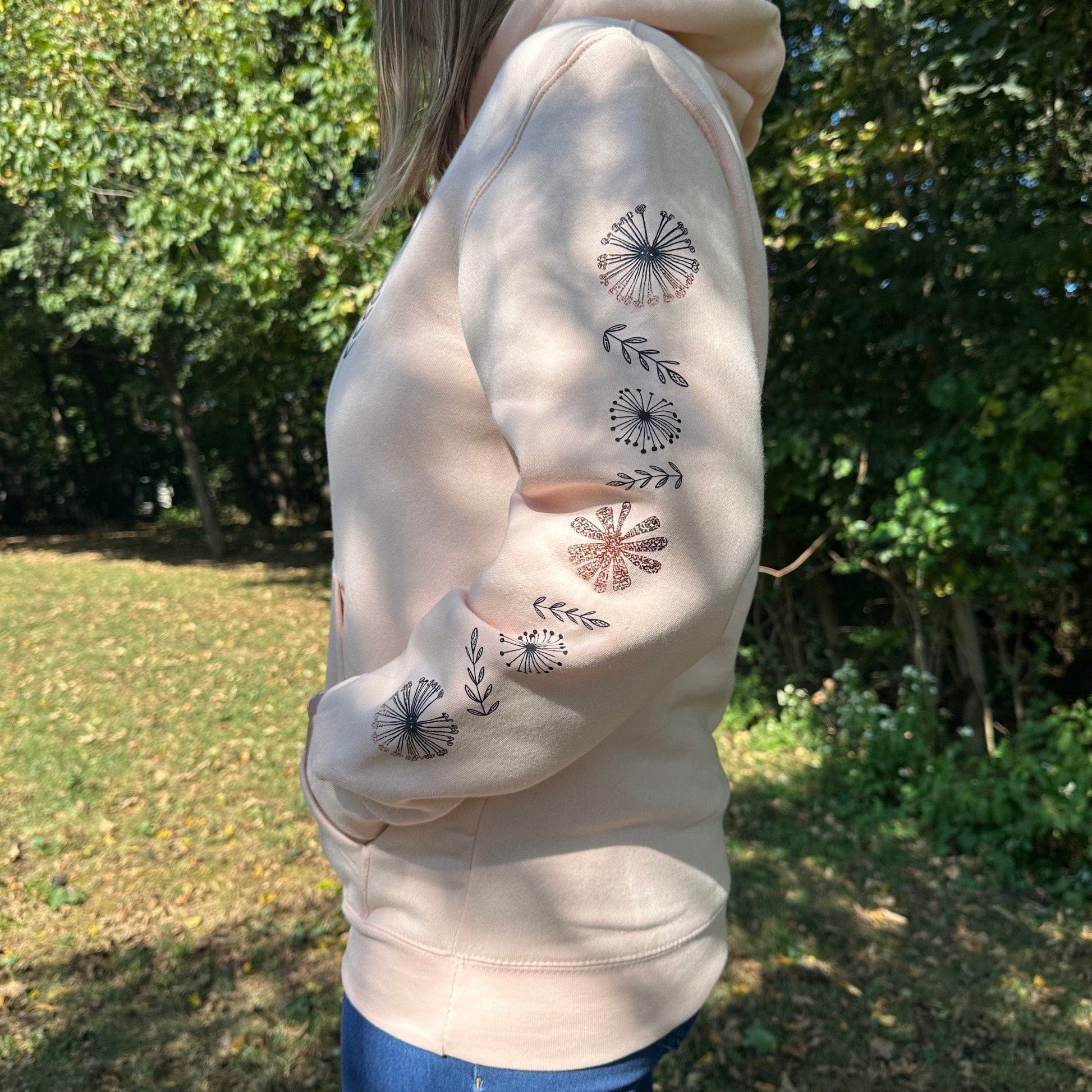 Woman standing in the woods wearing a pale pink hoodie that has a fun flower design down the sleeve.