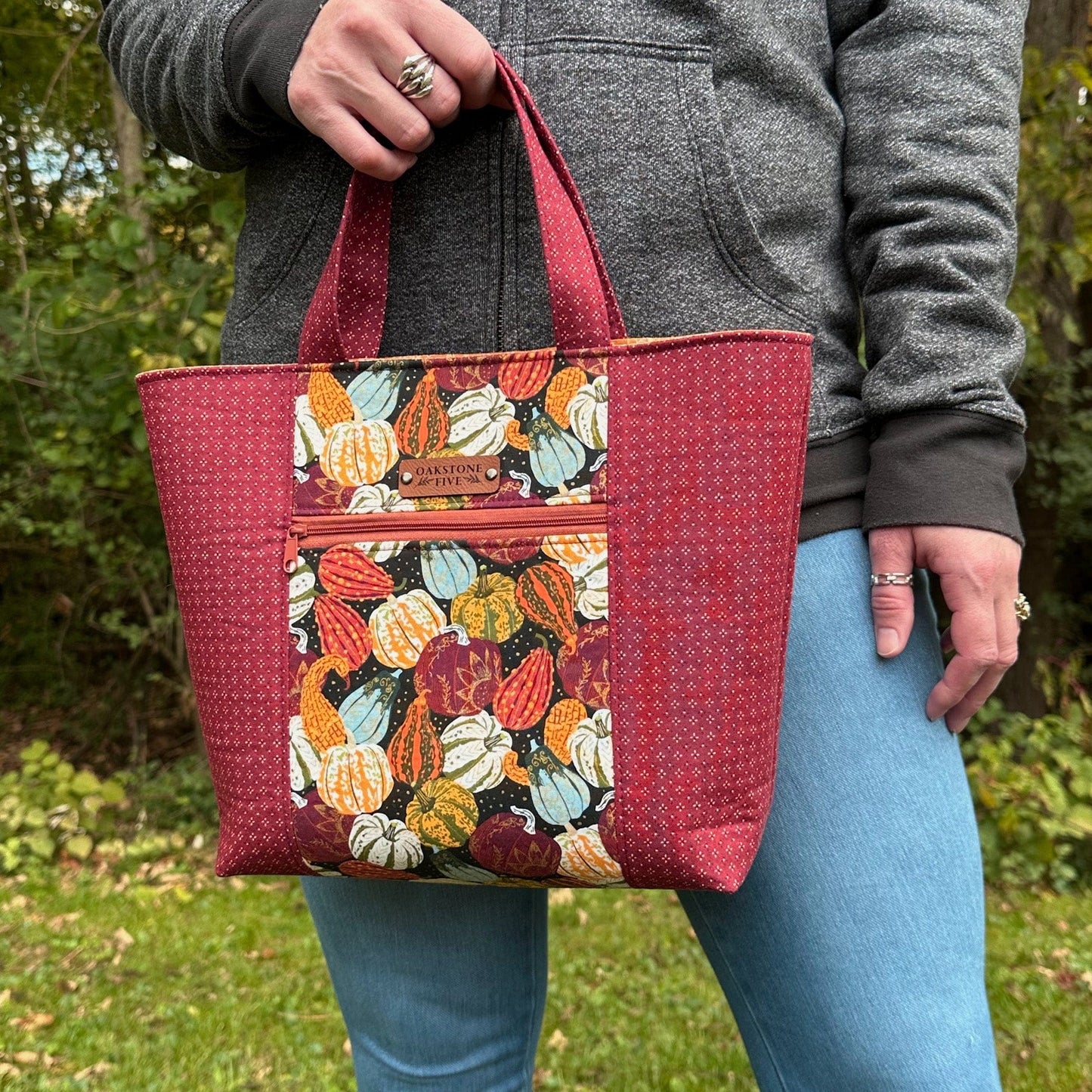 Woman standing in front of trees, holding a Fall tote bag with a gourd and pumpkin pattern. 