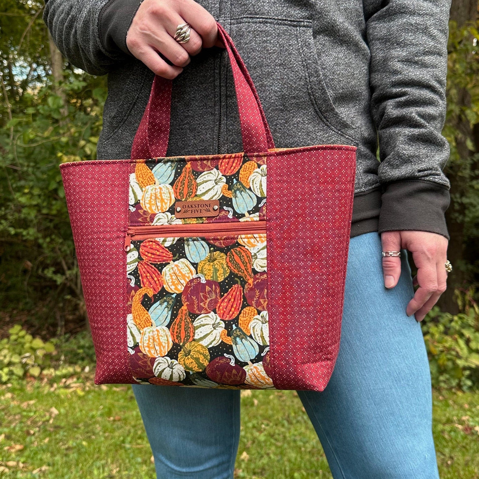 Woman standing in front of trees, holding a Fall tote bag with a gourd and pumpkin pattern. 