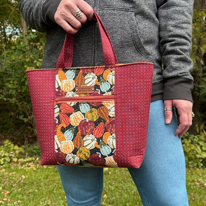 Woman standing in front of trees, holding a Fall tote bag with a gourd and pumpkin pattern. 